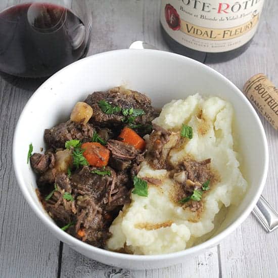 bowl of beef stew with side of mashed potatoes, served with a red wine.