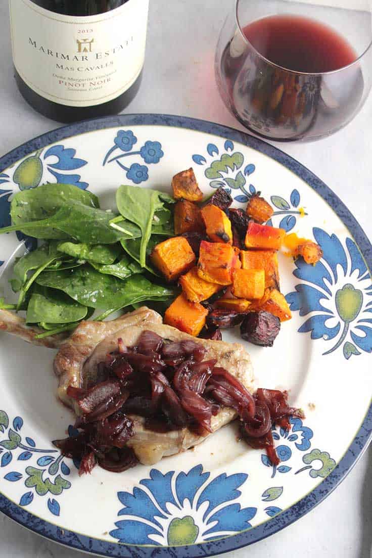 pork chop topped with pomegranate sauce, on a plate with vegetables, along with a glass of red wine.