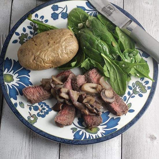 London broil steak topped with mushrooms on a plate with baked potato and salad.