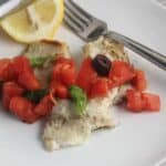 grilled tilapia topped with tomatoes and olives, with a lemon on the side.