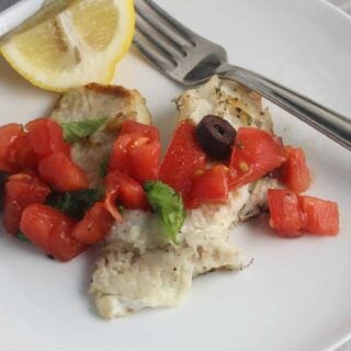 grilled tilapia topped with tomatoes and olives, with a lemon on the side.