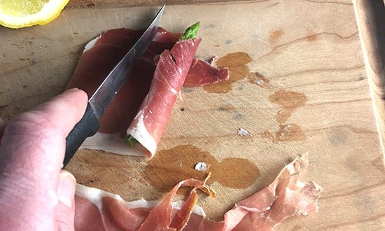 slicing a piece of extra prosciutto from a piece of asparagus wrapped with prosciutto.