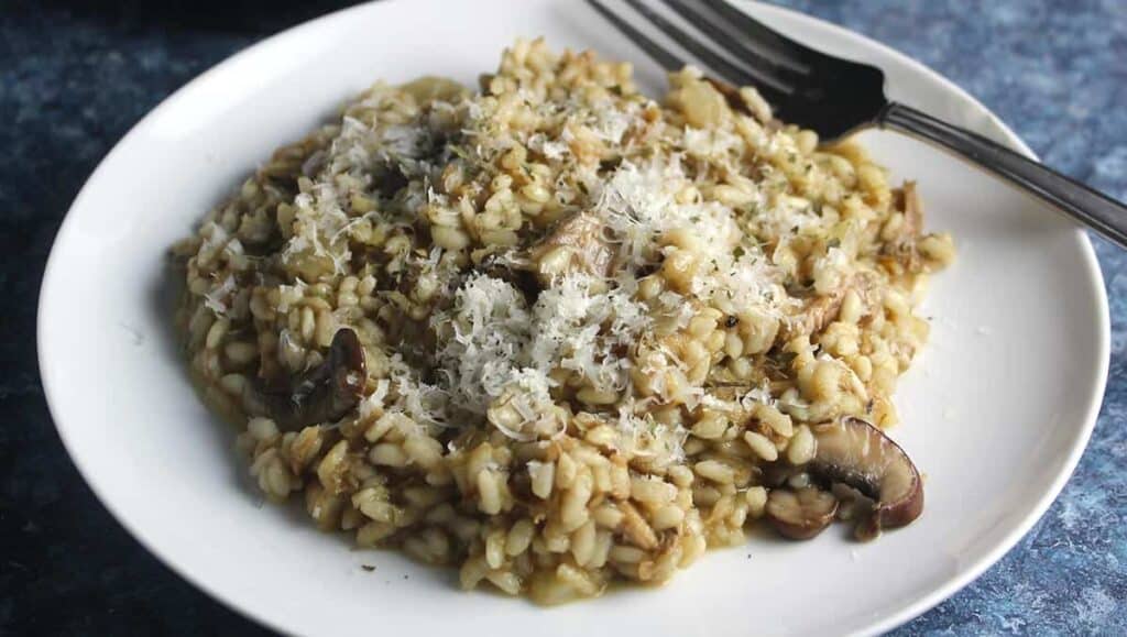 A white plate filled with mushroom risotto, topped with grated cheese. A fork is placed on the side of the plate on a blue textured surface. The risotto looks creamy and is mixed with pieces of mushroom.