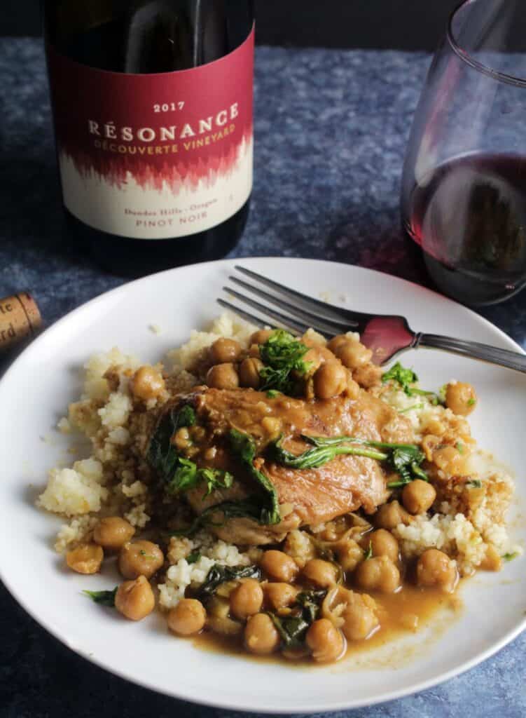 braised Moroccan chicken with chickpeas, served over couscous on a white plate. Bottle of Pinot Noir in the background.