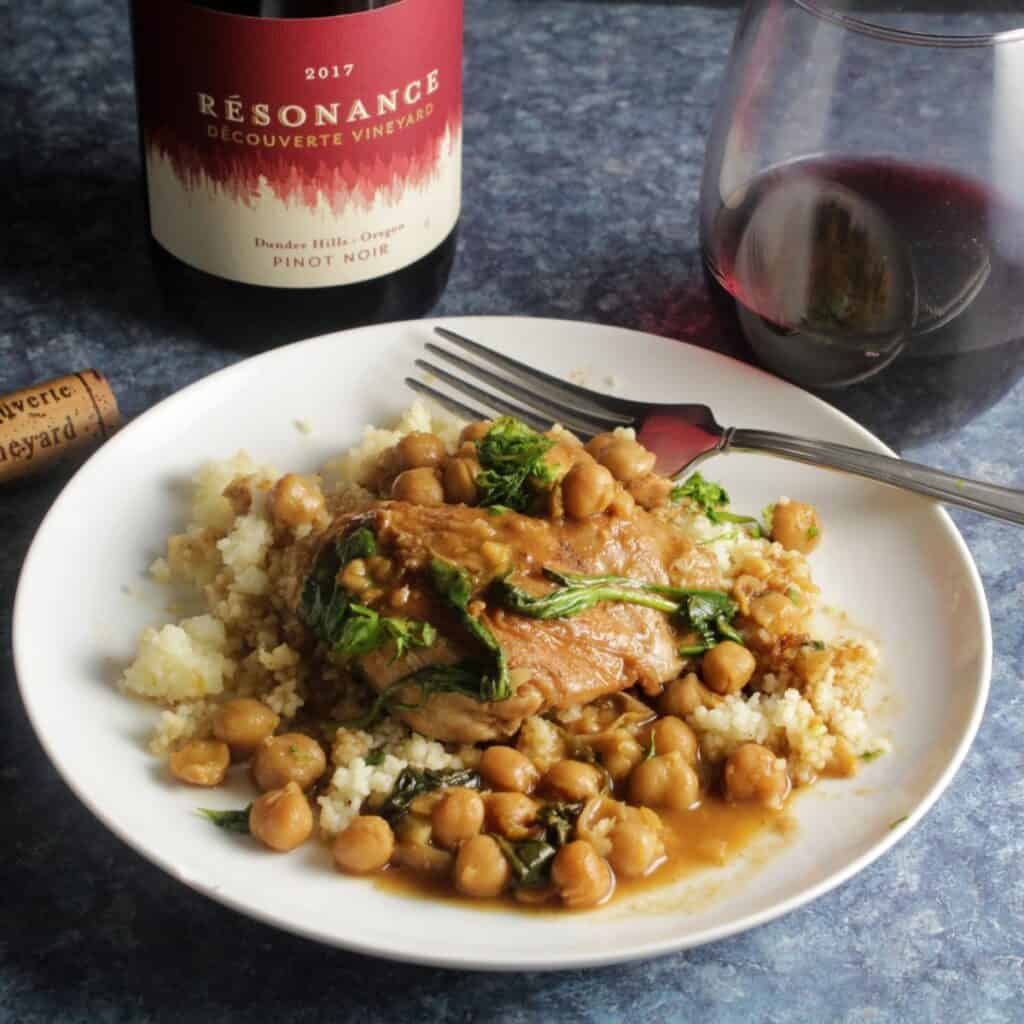 braised Moroccan chicken thighs with chickpeas, served over couscous with a bottle and glass of Pinot Noir red wine in the background.