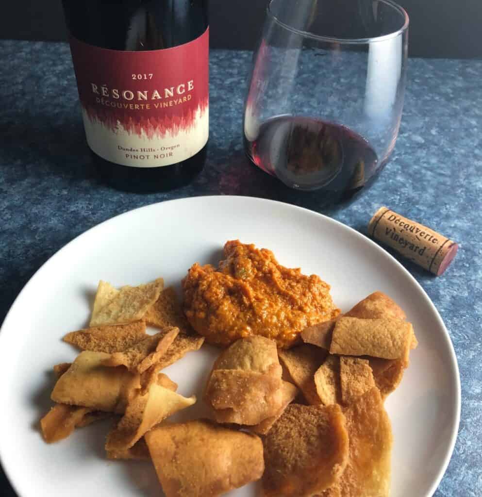 Muhammara red pepper dip on a plate with pita chips, and a red wine in the background.