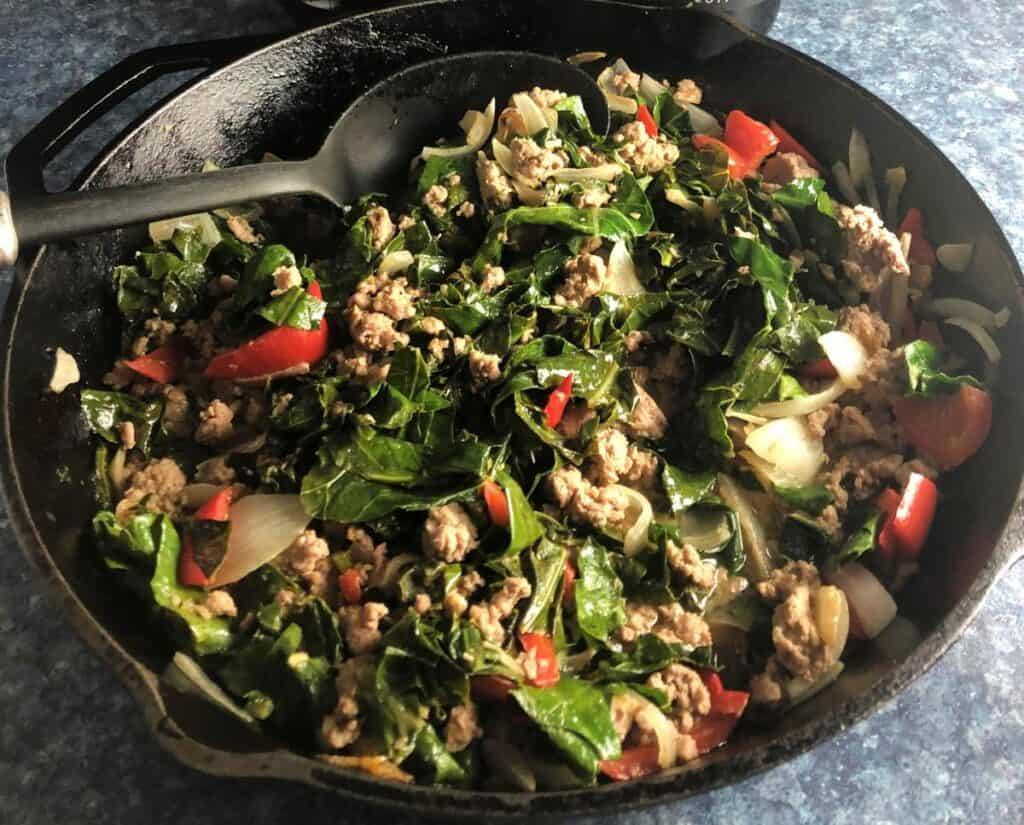 stirring ground pork stir fry with collard greens in a black skillet.