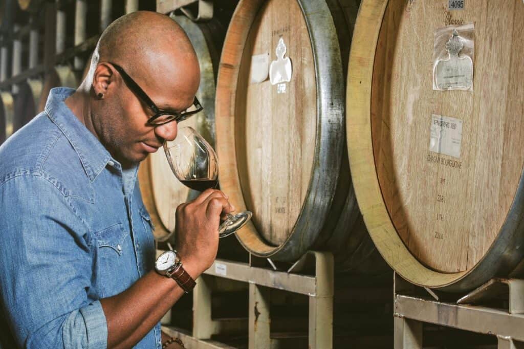 André Hueston Mack tasting wine with wine barrels behind him.