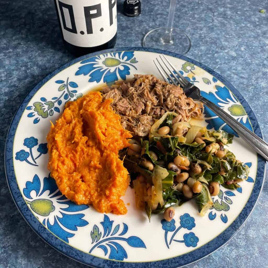 black-eyed peas collard greens, pulled pork and mashed sweet potatoes on a plate with blue flowers on the edge.