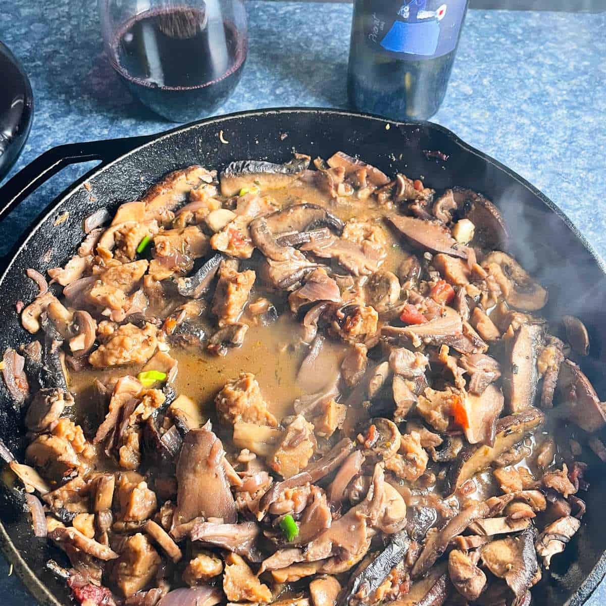 skillet with mushrooms cooked in sauce with steam rising. Red wine in the background.
