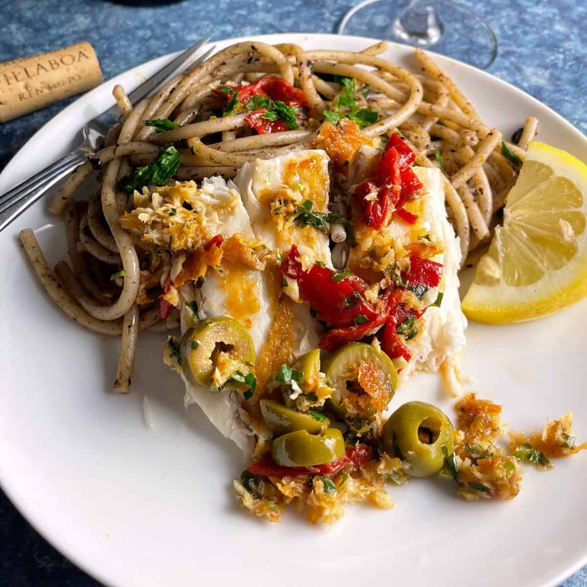 pan seared sea bass topped with a sauce including olives, red peppers and parsley. Served with pasta and a slice of lemon.