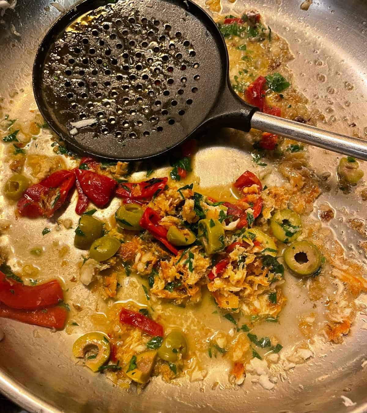stirring a sauce in a silver skillet, with olives, red peppers and parsley
