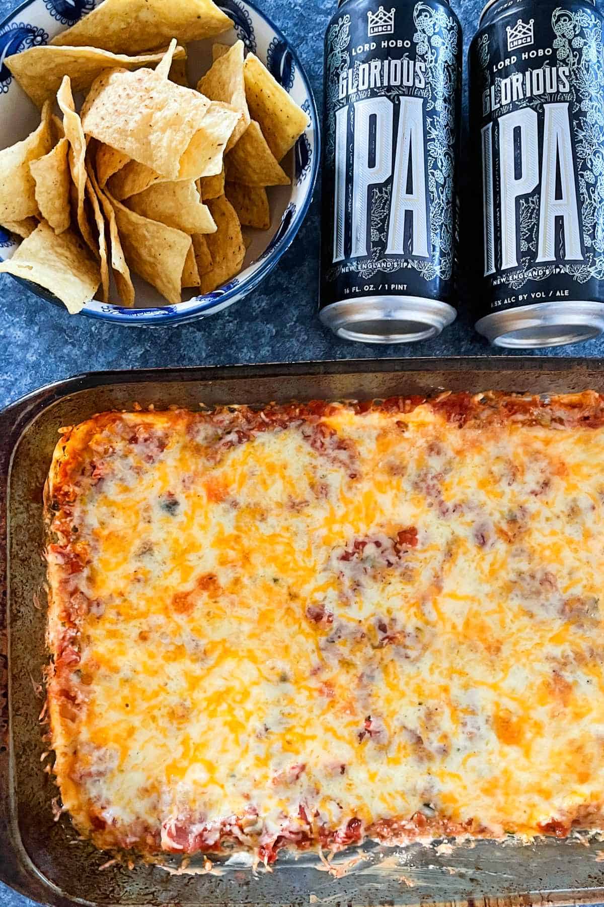 Mexican dip with melted cheese in a baking dish, with cans of Glorious IPA beer above it.