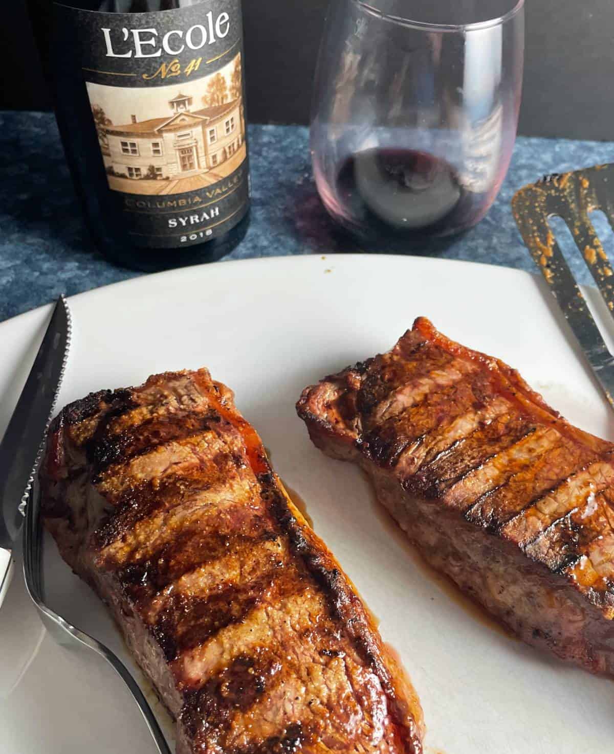 grilled NY strip steaks on a white platter, served with a Syrah red wine in the background.
