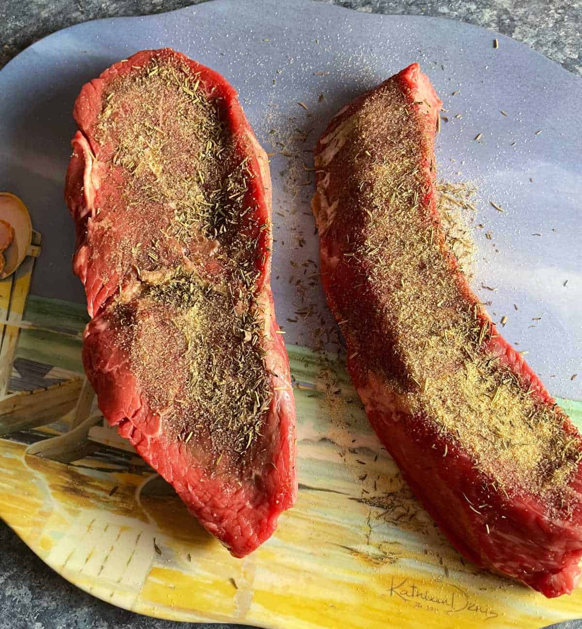 two raw sirloin steaks with dried spice rub on a platter, ready for grilling.