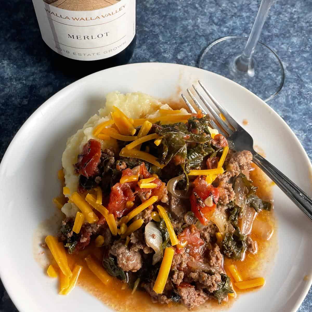 ground beef and kale topped with cheddar cheese, served over mashed potatoes. Red Merlot wine in the background.