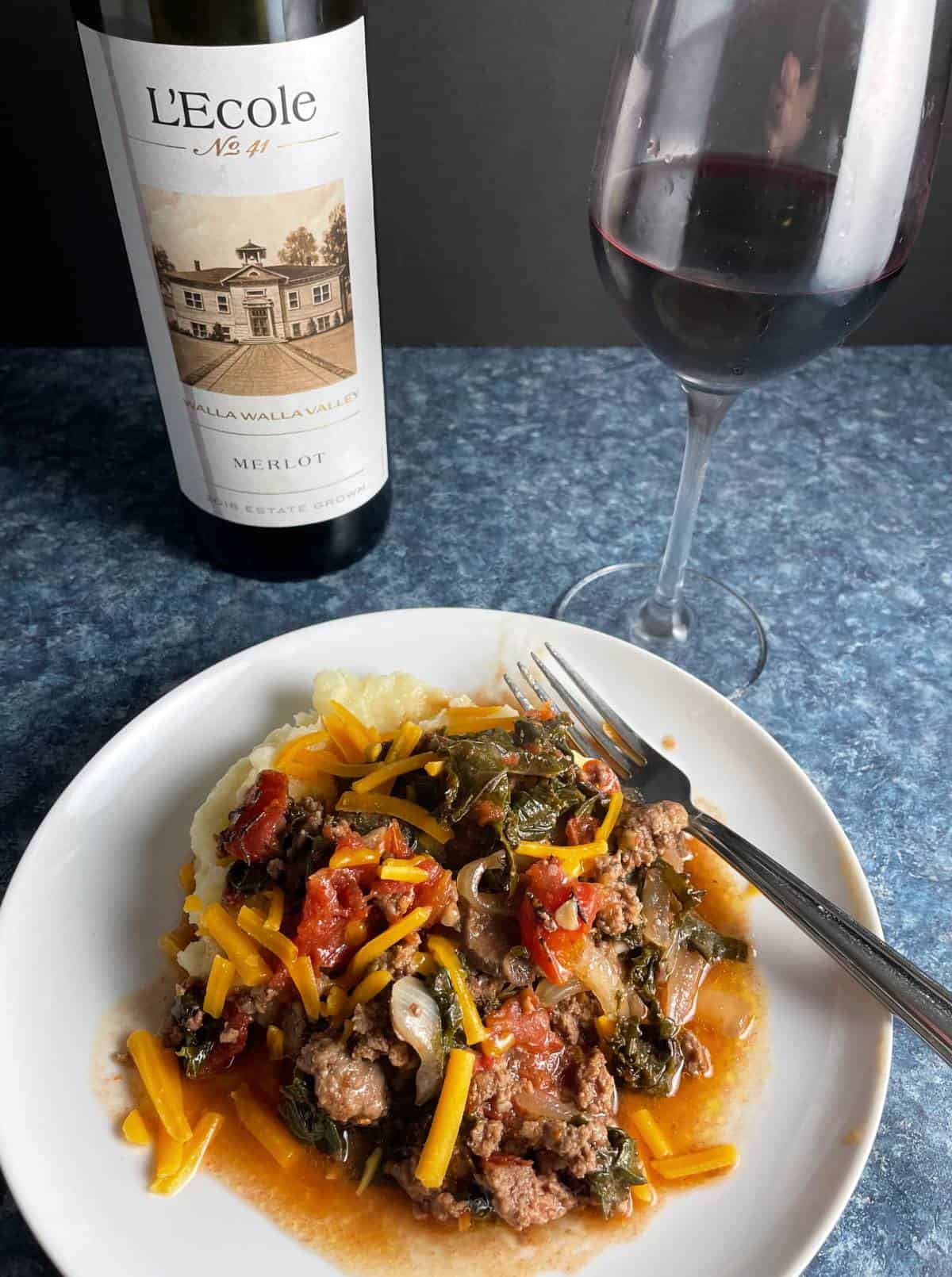 ground beef and kale topped with cheddar cheese, served over mashed potatoes on a white plate. Red wine in the background. 