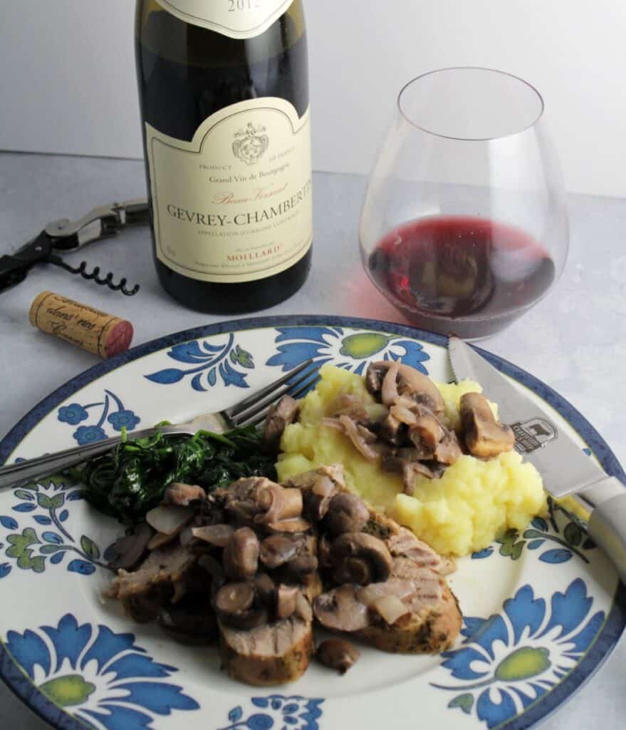 pork tenderloin topped with mushrooms, served with mashed potatoes and greens. Bottle and glass of red Burgundy wine in background.