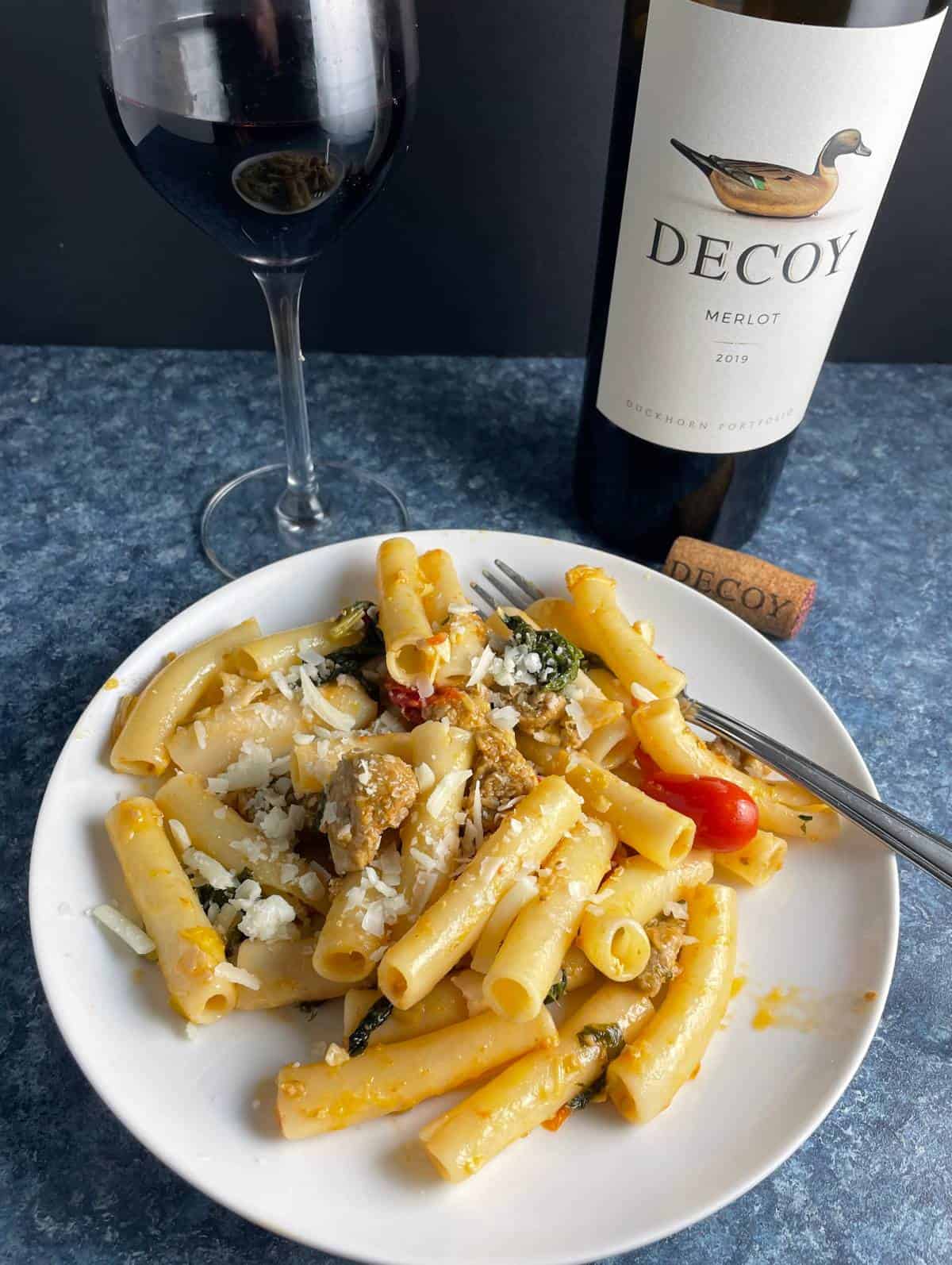 ziti pasta with sausage and greens on a white plate, served with a Merlot red wine.