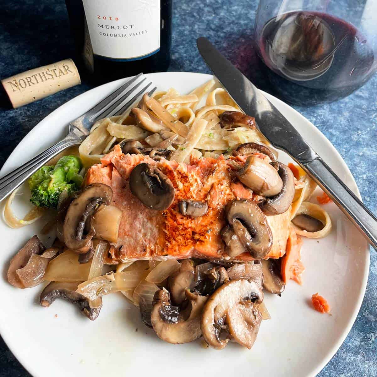 salmon topped with mushrooms, served with pasta and red wine in the background.