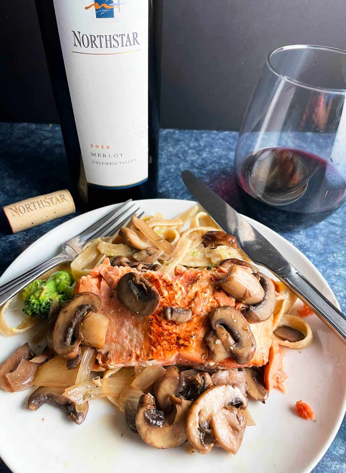 seared salmon topped with mushrooms, served with fettuccine. Glass and bottle of Merlot red wine in the background.