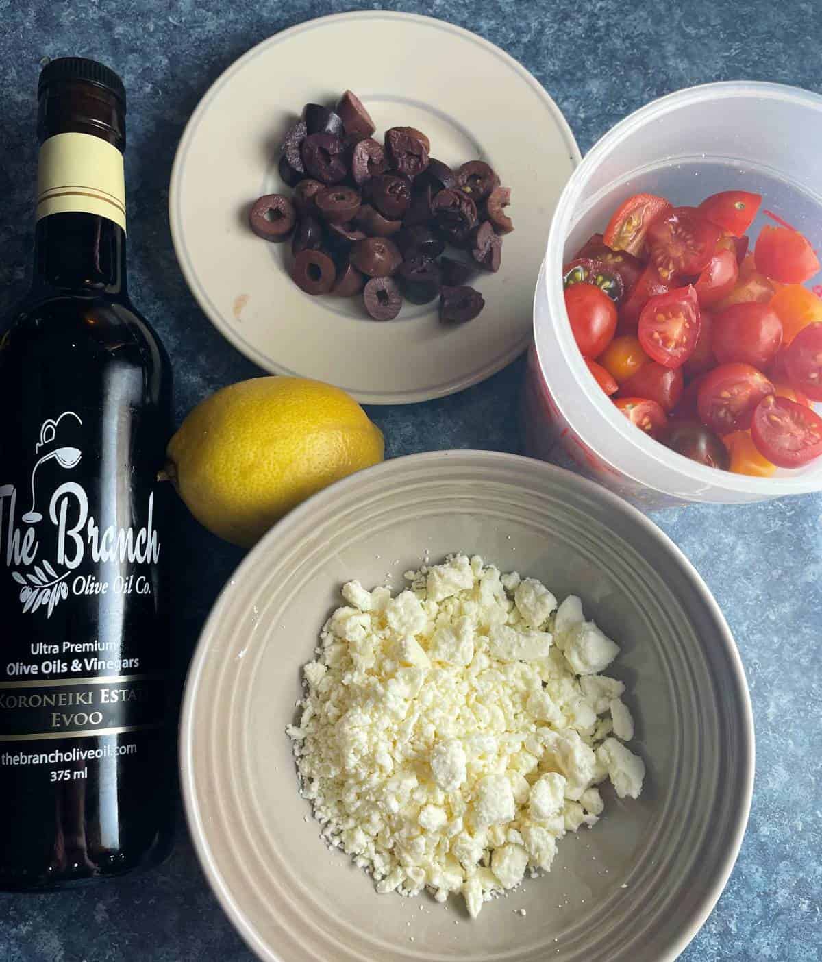 ingredients for a greek pasta recipe on a blue counter top -- feta cheese, extra virgin olive oil, lemon and sliced olives.