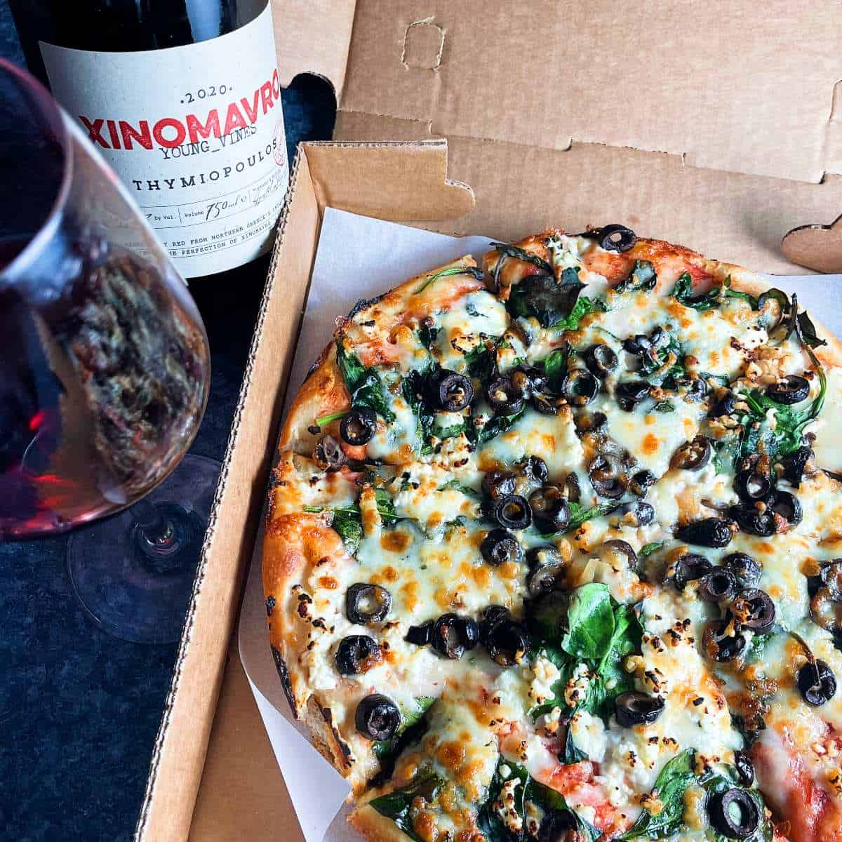 pizza in an opened takeout box, topped with feta, spinach, garlic and olives. Greek red wine alongside the pizza.