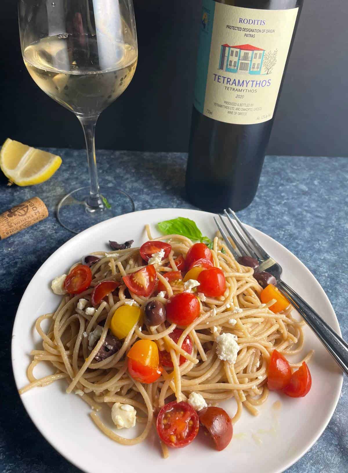 Greek flavored spaghetti served on a white plate, topped with tomatoes and feta cheese, served with white wine. 