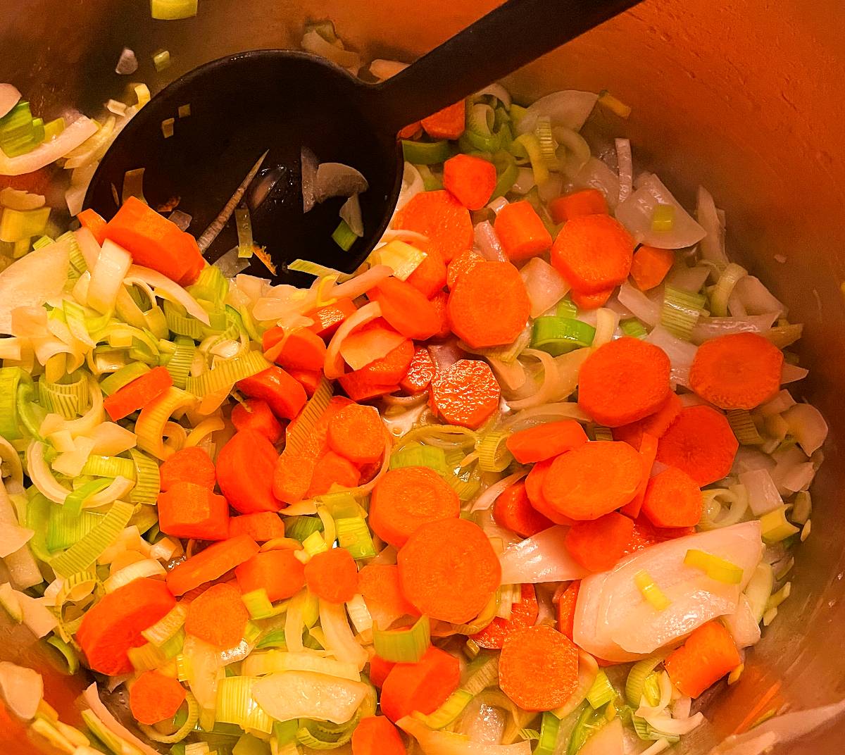 adding carrots to a soup pot with leeks and onions.