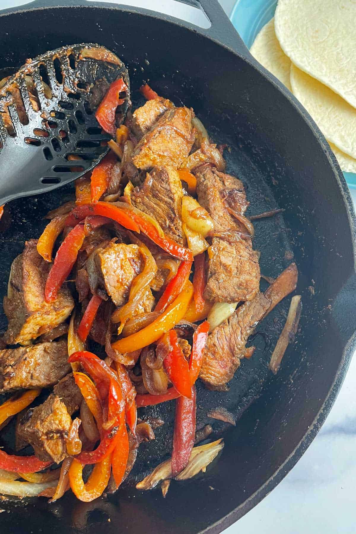slices of steak with onions and peppers in a black skillet. 