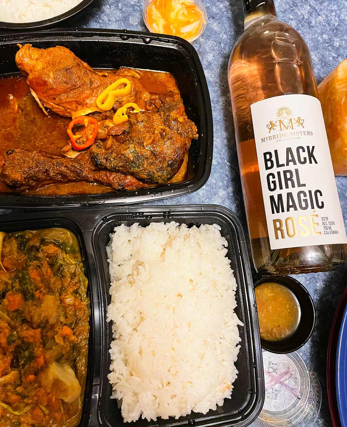 Haitian chicken, rice, and vegetables in takeout containers alongside a bottle of Black Girl Magic Rosé wine.