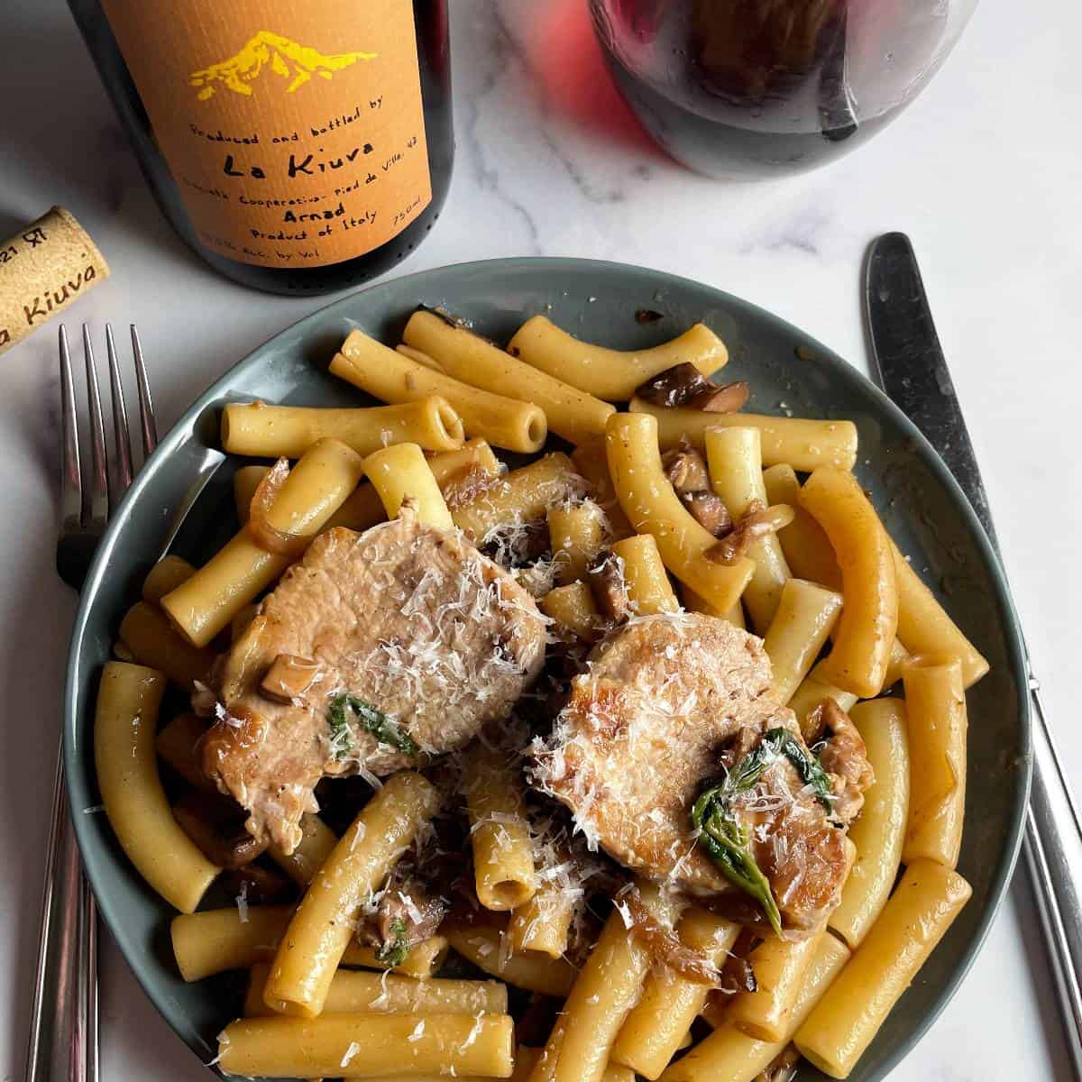 ziti pasta served with pork tenderloin, caramelized onions, basil and parmesan cheese. glass of red wine in the foreground.