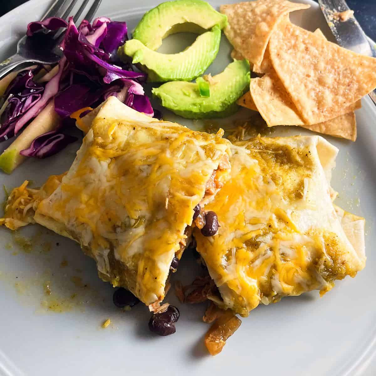 chicken and black bean enchiladas served on a plate with red cabbage, avocado slices and tortilla chips.