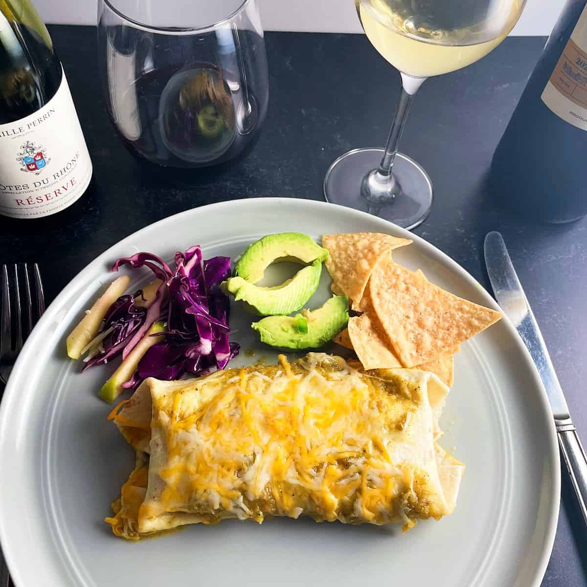 chicken and black bean enchiladas on a plate with red cabbage slaw and avocado slices. Red and white wine in the background.