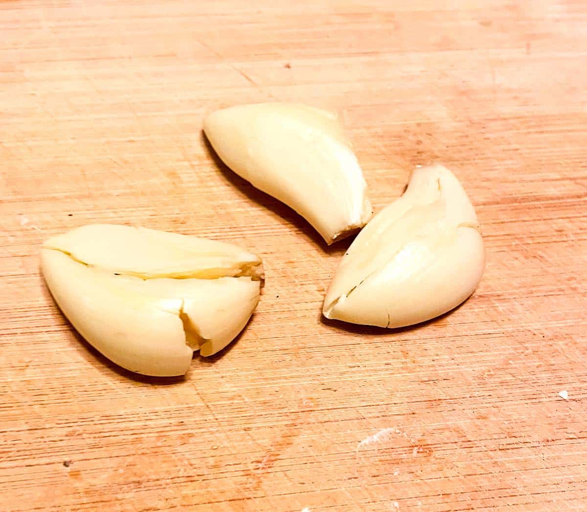 3 peeled garlic cloves on a wooden cutting board.