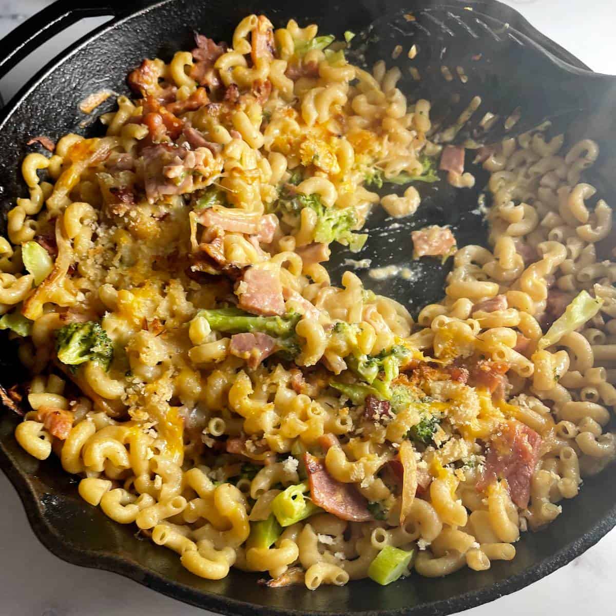 ham, cheese and pasta baked in a large black skillet. photo shows a spoon scooping out a serving.