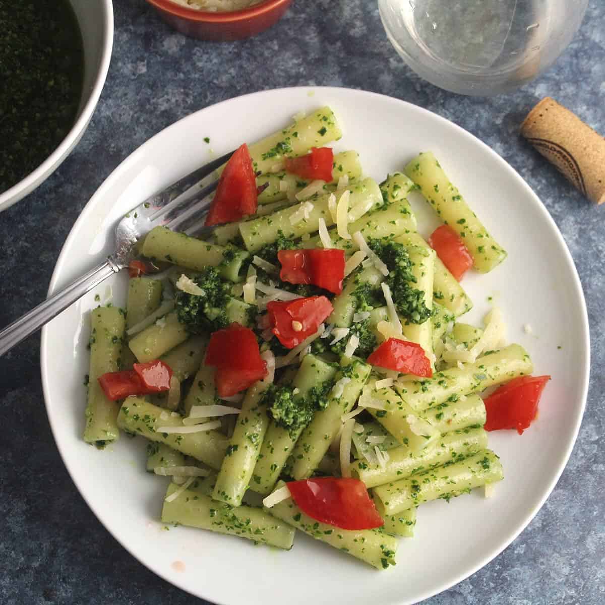 ziti tossed with kale pesto and topped with tomatoes. served on a while plate with a fork.