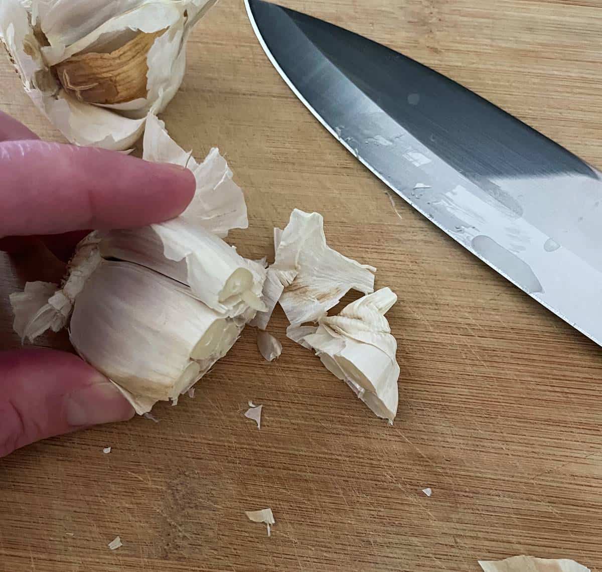 cutting the root end of garlic off using a large knife on a wooden cutting board. 