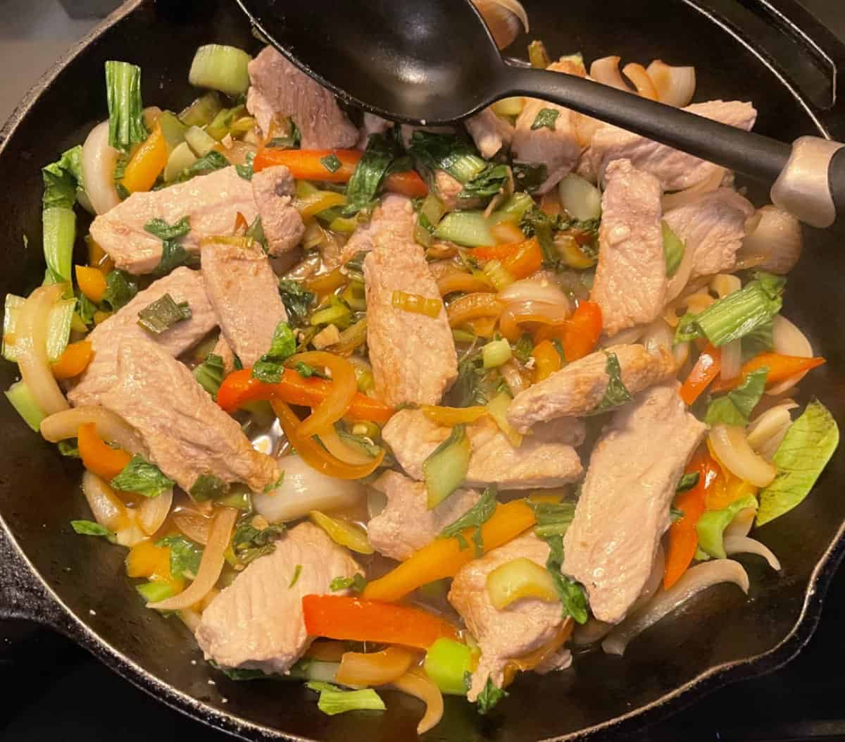 Cooked pork, sliced white onion, red pepper, orange pepper, and bok choy prepared in a black frying pan.