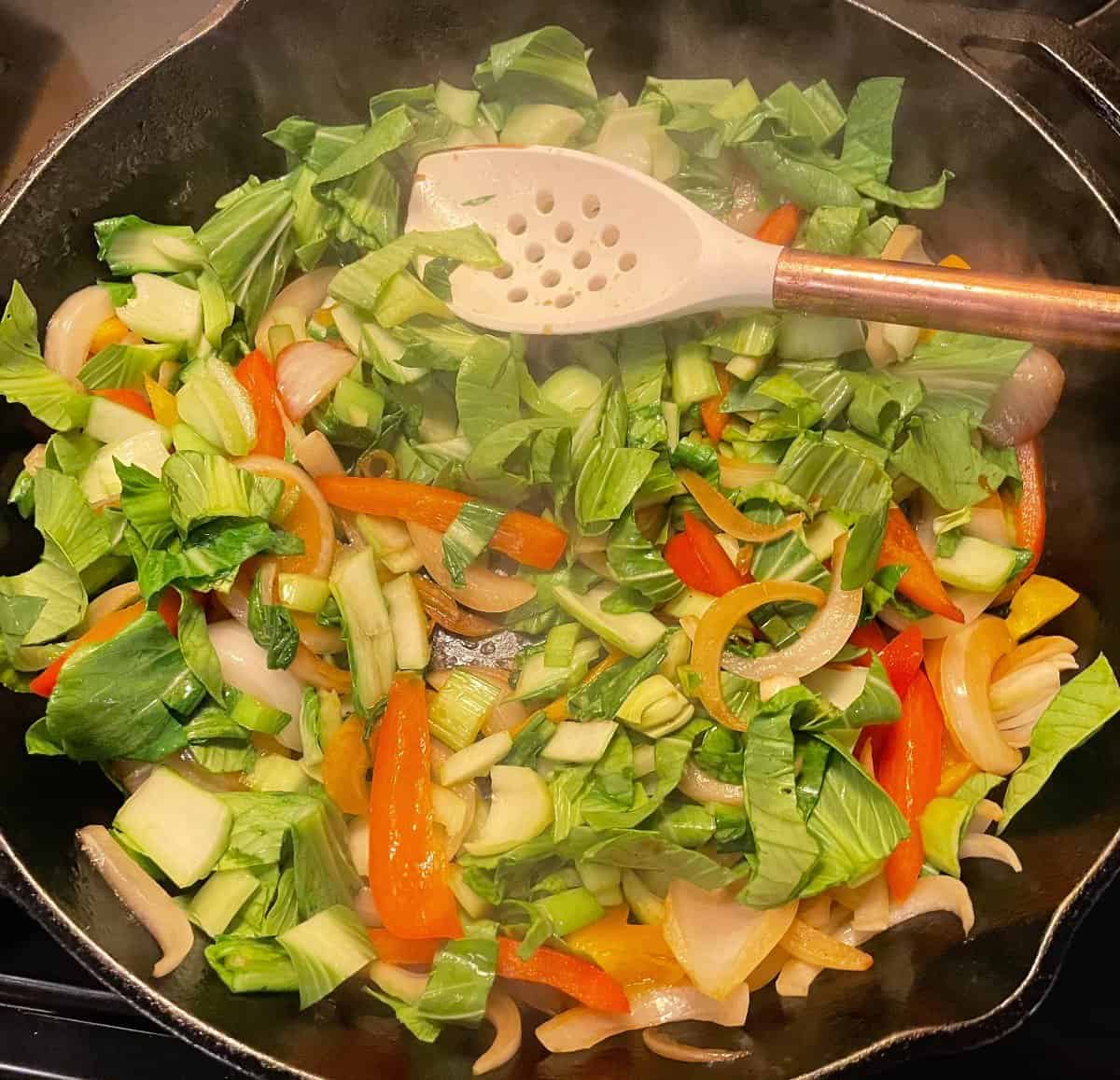 Chopped bok choy, sliced red peppers, orange peppers, and white onions in a black frying pan.