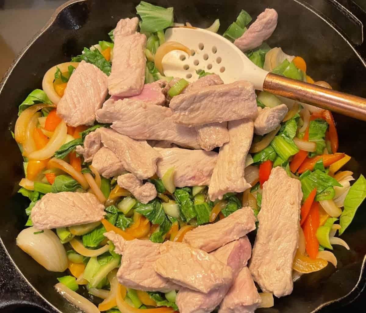 Sliced pork, white onions, red peppers, orange peppers, and bok choy sautéing in a black frying pan.