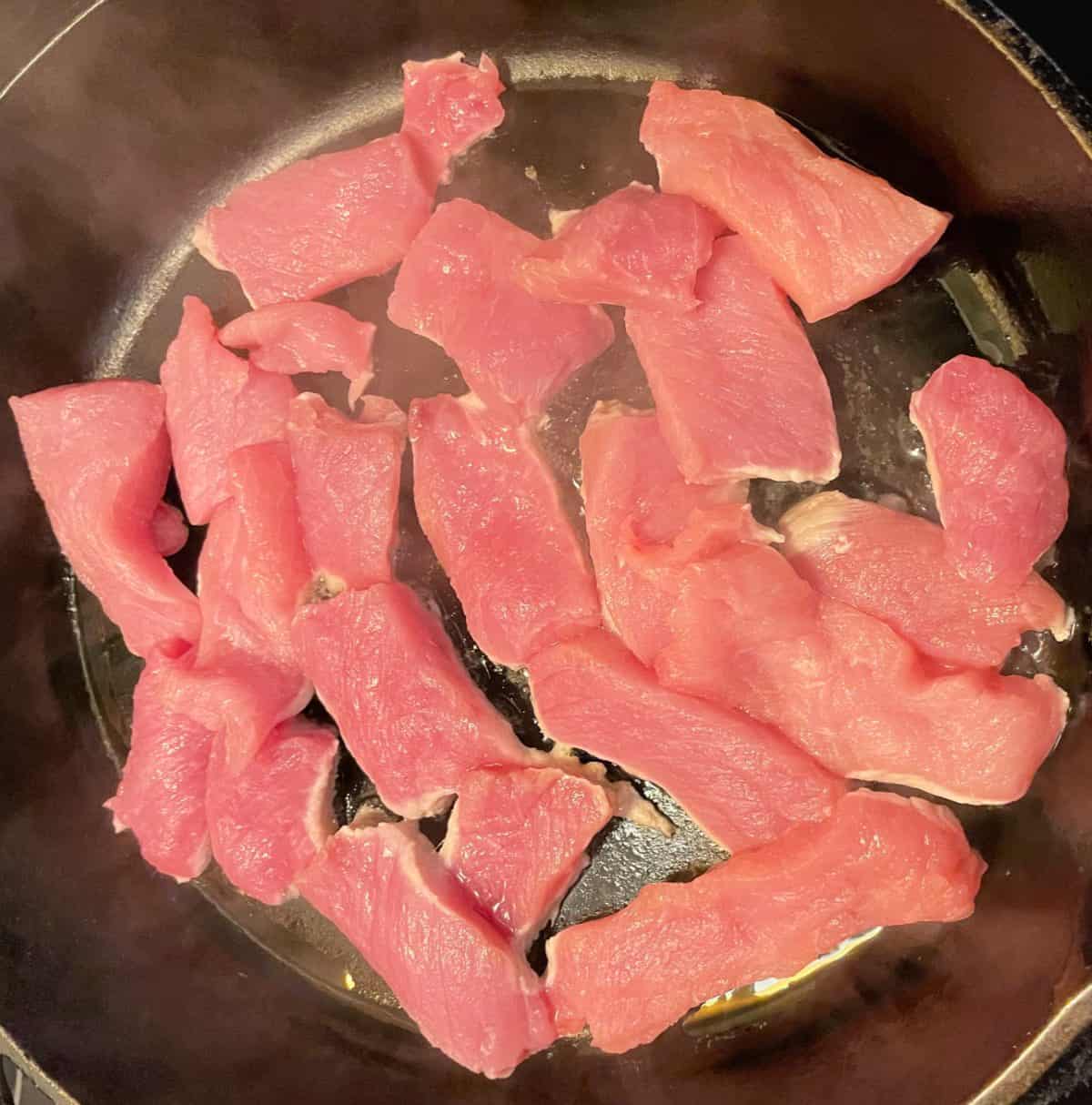 Pork chop pieces cooking in a black frying pan. 