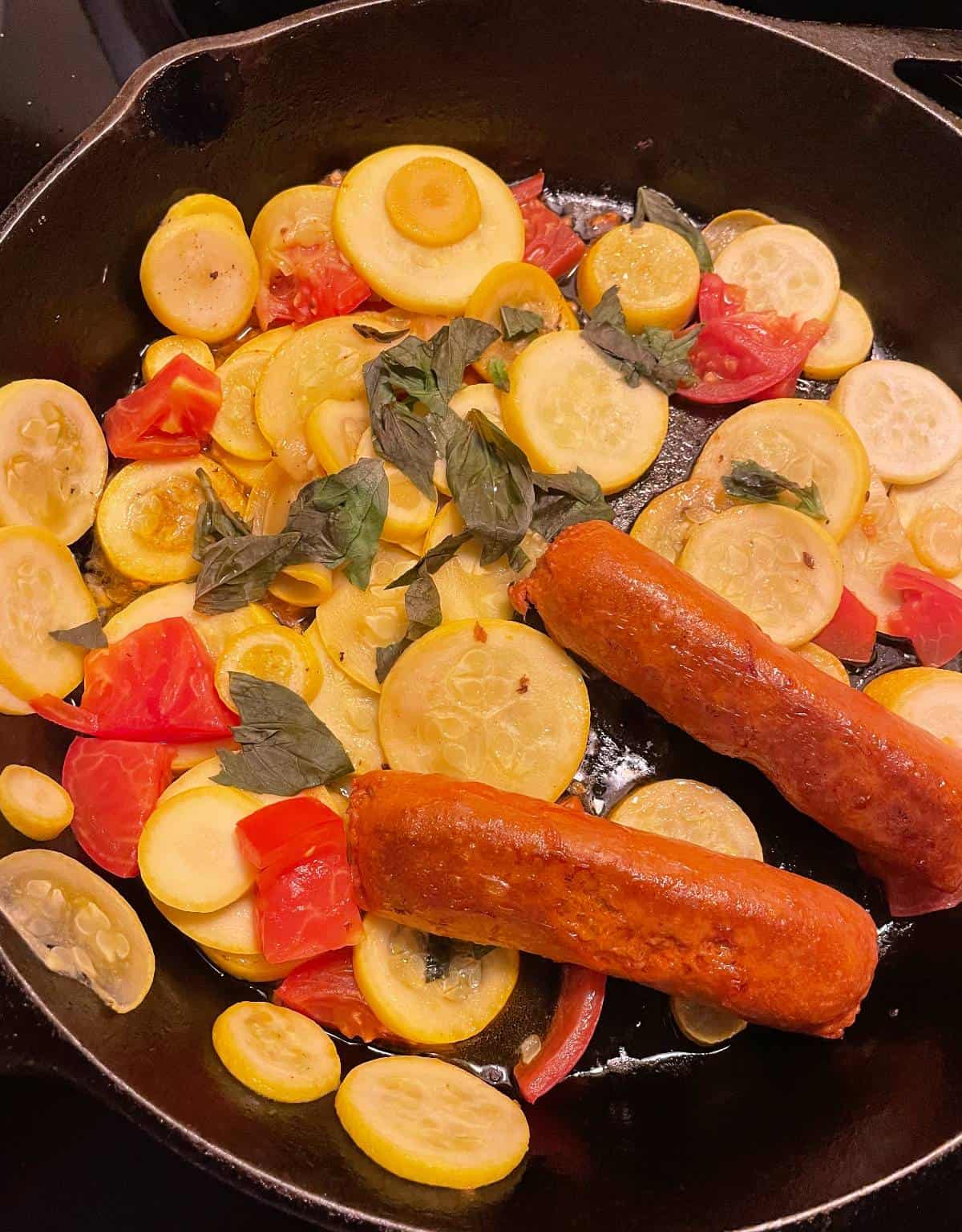 sliced yellow summer squash in a black skillet along with tomatoes and vegetarian sausage.