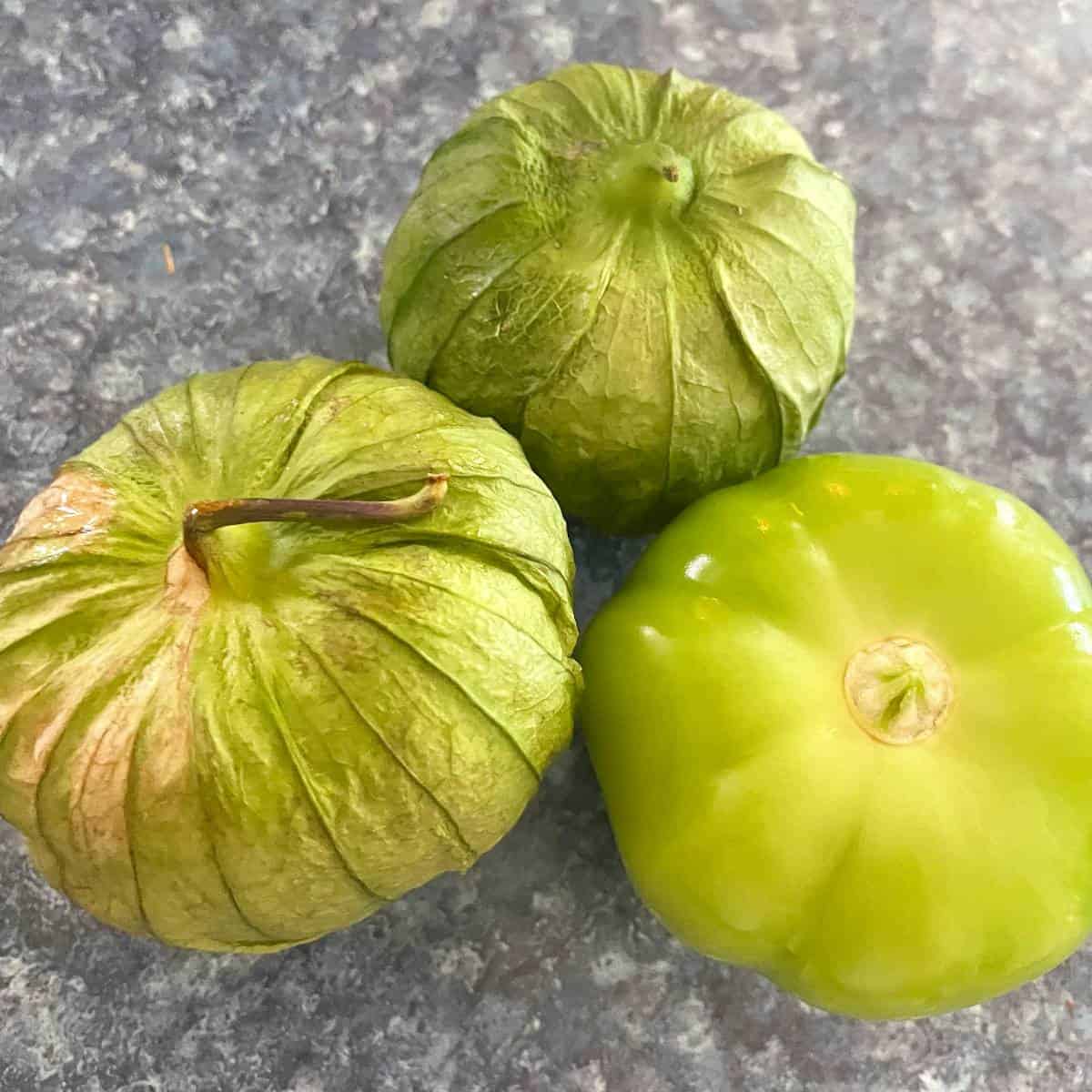 3 tomatillos on a blue counter top. 2 of the tomatillos have their husk on, one has it removed.
