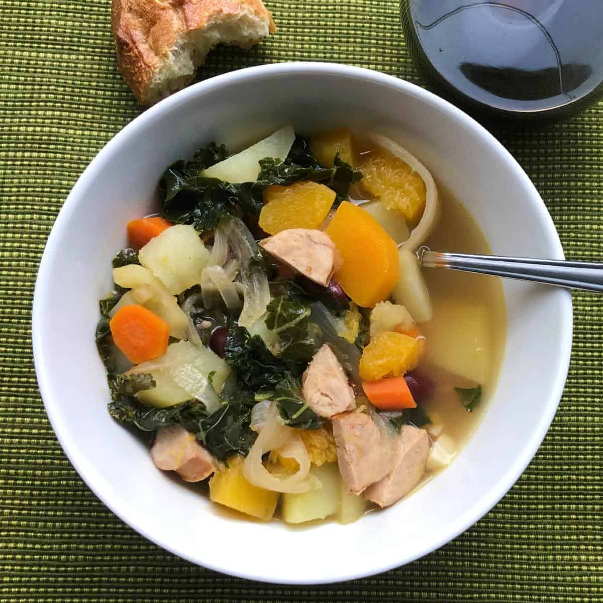 Portuguese Kale Soup, with kale, potatoes carrots and sausages, served in a white bowl on a green placemat. 