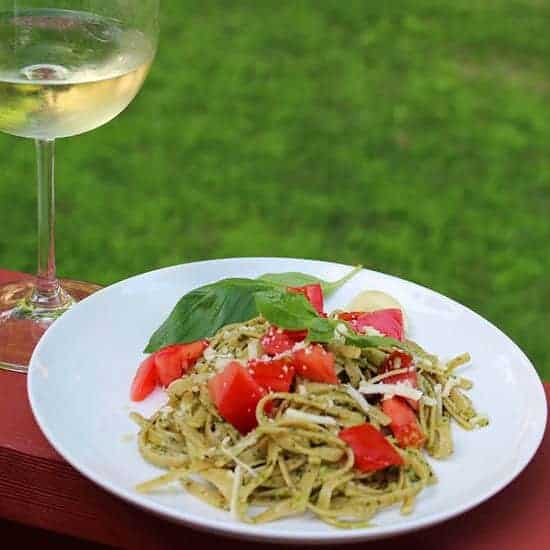 linguine with pesto sauce served on a white plate, topped with pieces of tomato with a white wine in the background. 