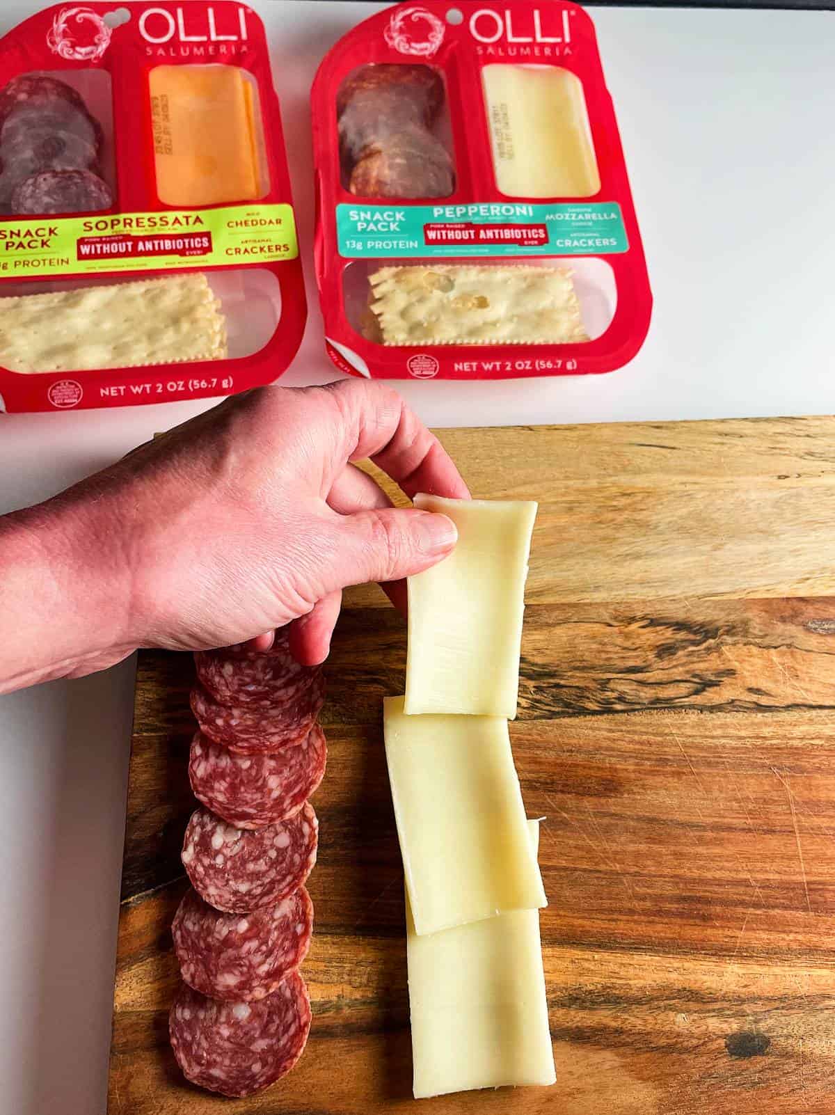 a hand arranging white cheese slices alongside salami slices on a wooden board. 