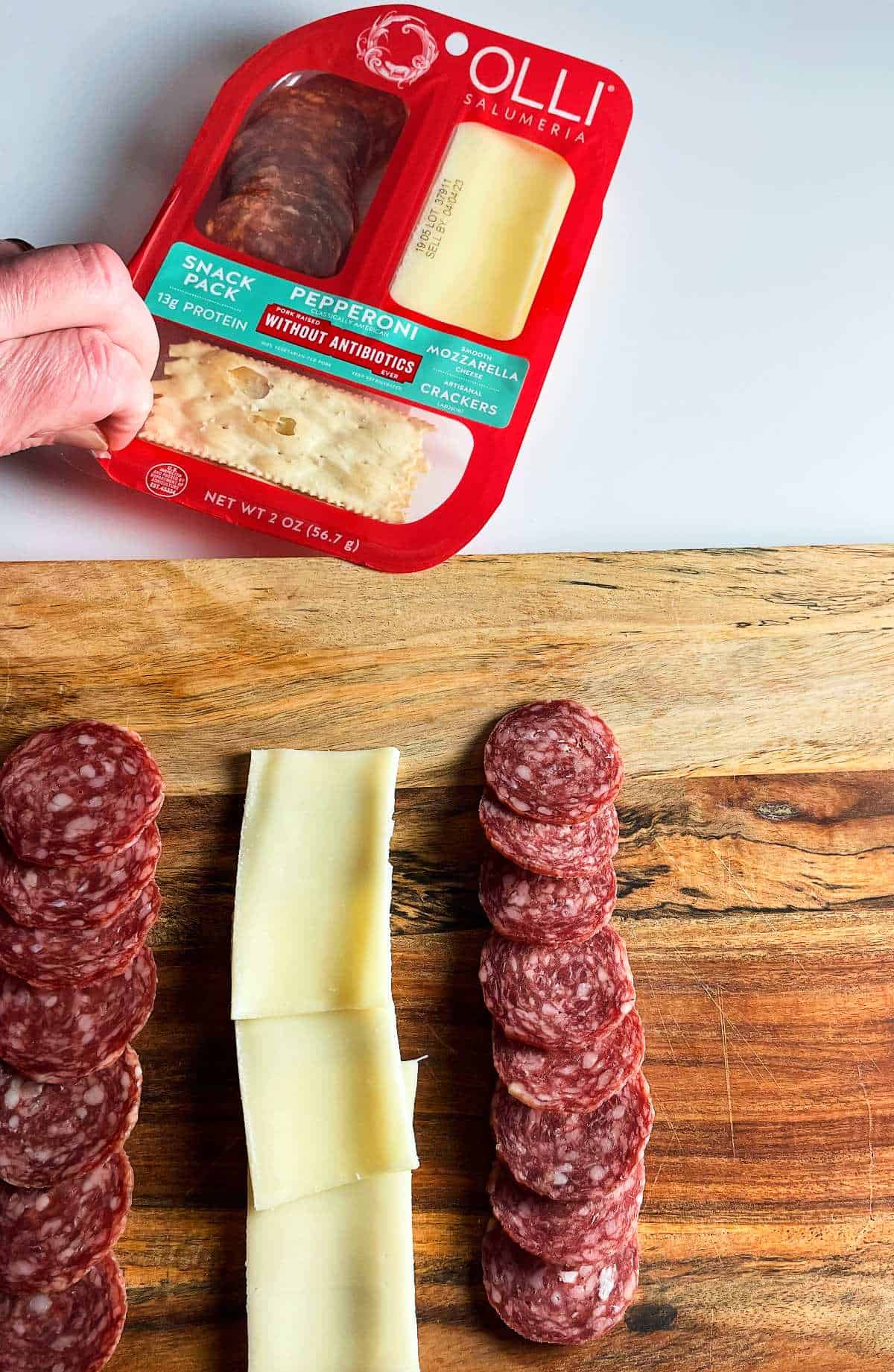 arranging salami and cheese on a wooden cutting board. Olli Salumeria Snack Pack above the cutting board. 