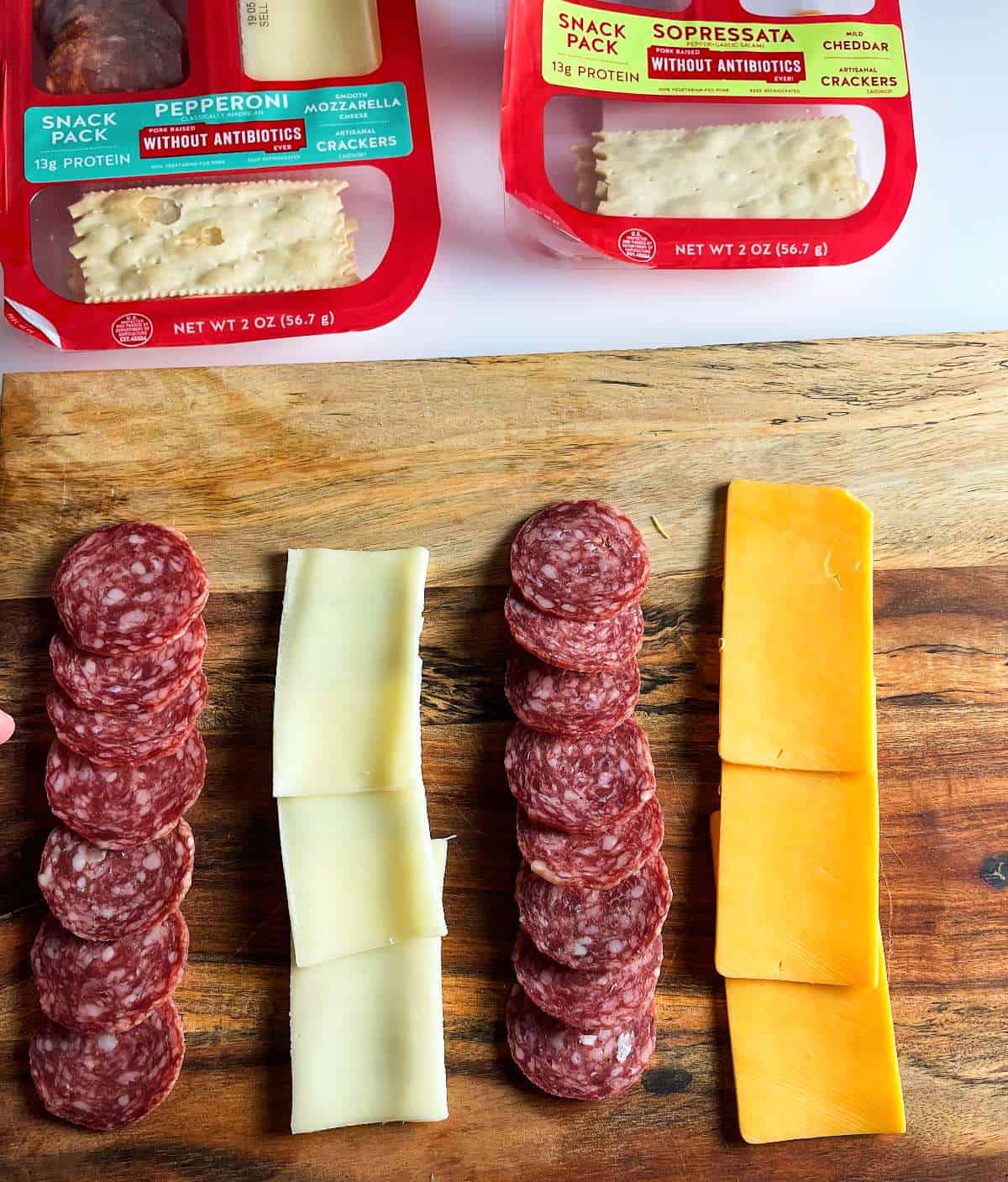 cheese and salami arranged on a wooden board, shown from above. Olli Salumeria Snack Packs are next to the board. 