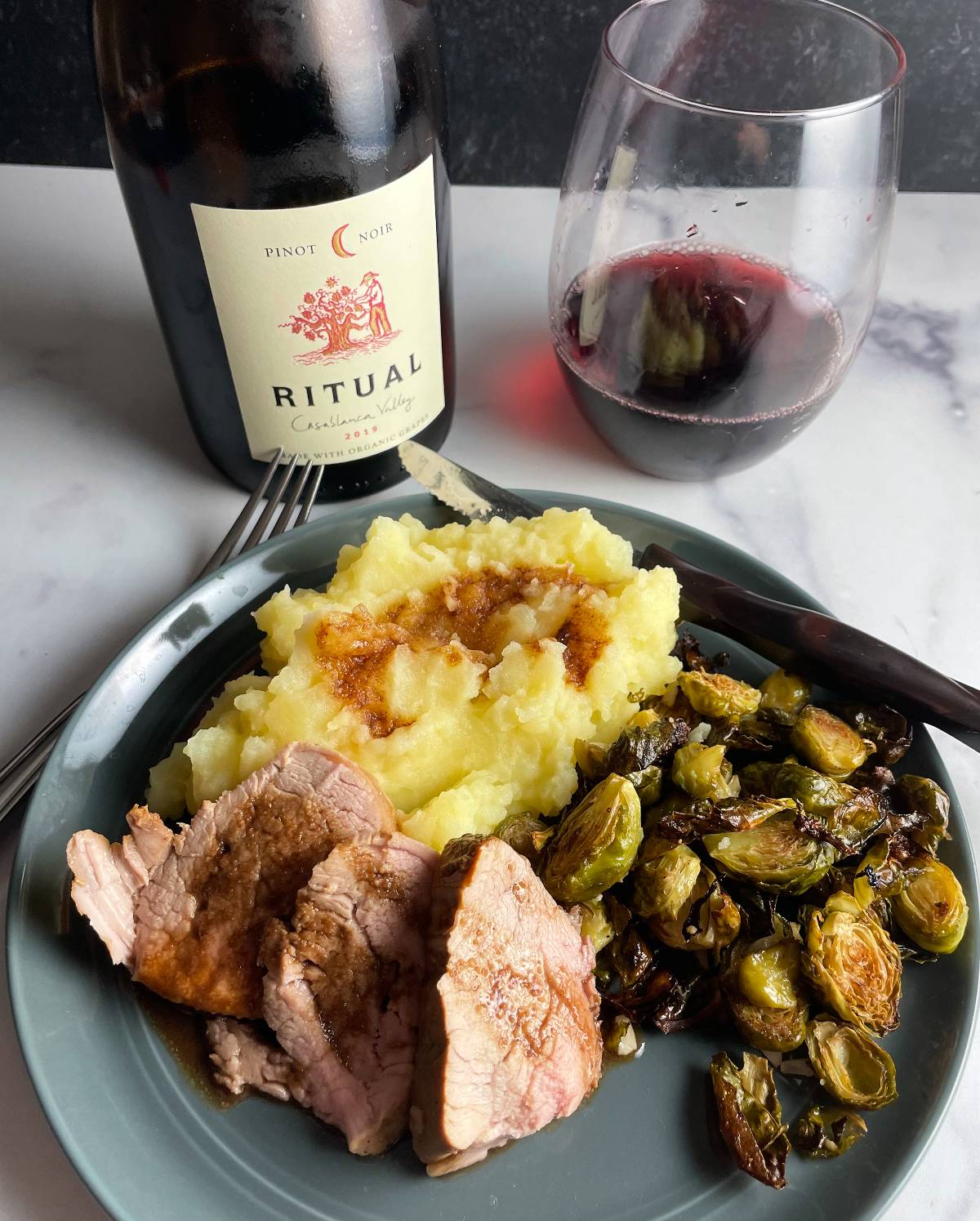 Slices of pork tenderloin along with mashed potatoes and roasted Brussels sprouts, with a Pinot Noir in the background.